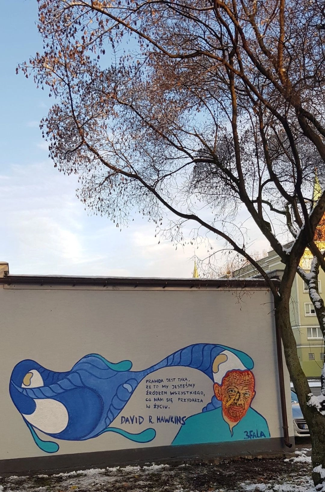 Kolorowy mural z cytatem Davida R. Hawkinsa i stylizowaną postacią na ścianie budynku w Warszawie, zimowy krajobraz wokół.