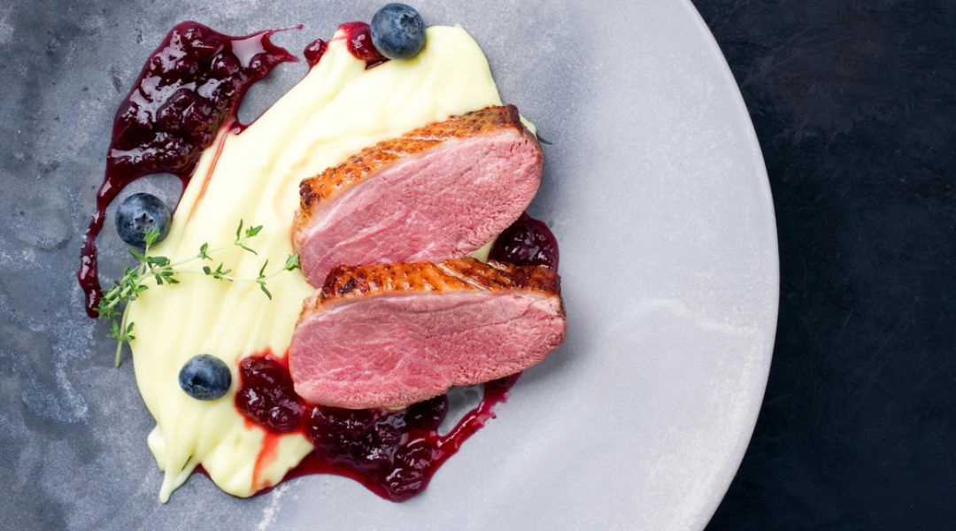 Plastry piersi z gęsi na kremowym puree z sosem żurawinowym i borówkami na szarym talerzu