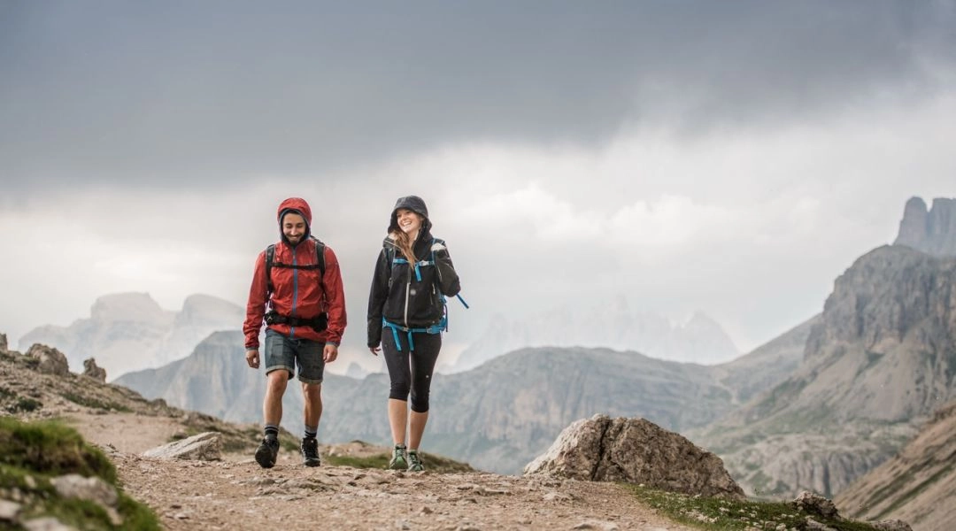 Dwoje turystów w kurtkach przeciwdeszczowych idzie kamienistym szlakiem w Dolomitach podczas deszczowej pogody.