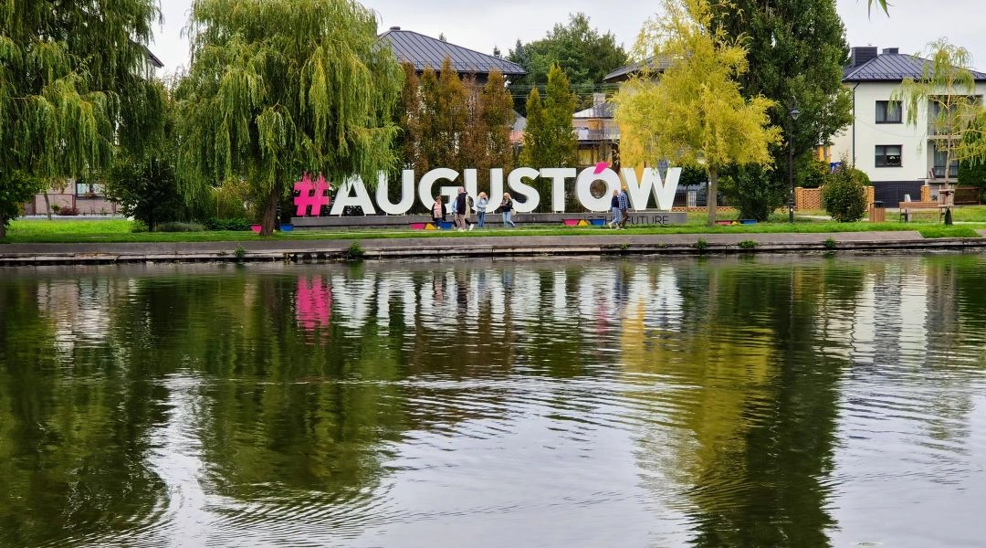 Duży napis #AUGUSTÓW ustawiony nad wodą, otoczony zielonymi drzewami i odbijający się w tafli.