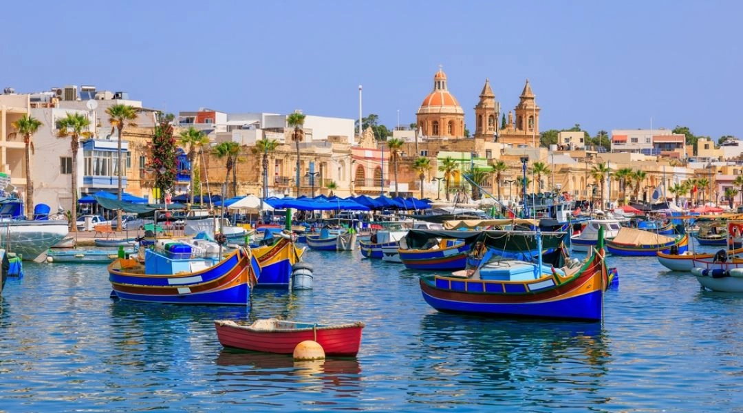 Kolorowe tradycyjne łodzie luzzu w porcie Marsaxlokk na Malcie, nadbrzeże i kościół parafialny w tle