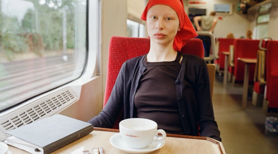 Portret młodej kobiety siedzącej w pociągu przy stoliku, w czerwonej chuście i czarnym swetrze, patrzącej w obiektyw.