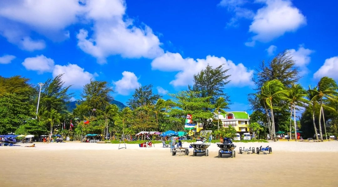 Szeroka piaszczysta plaża na Phuket z palmami, parasolami, ludźmi i skuterami wodnymi stojącymi na brzegu