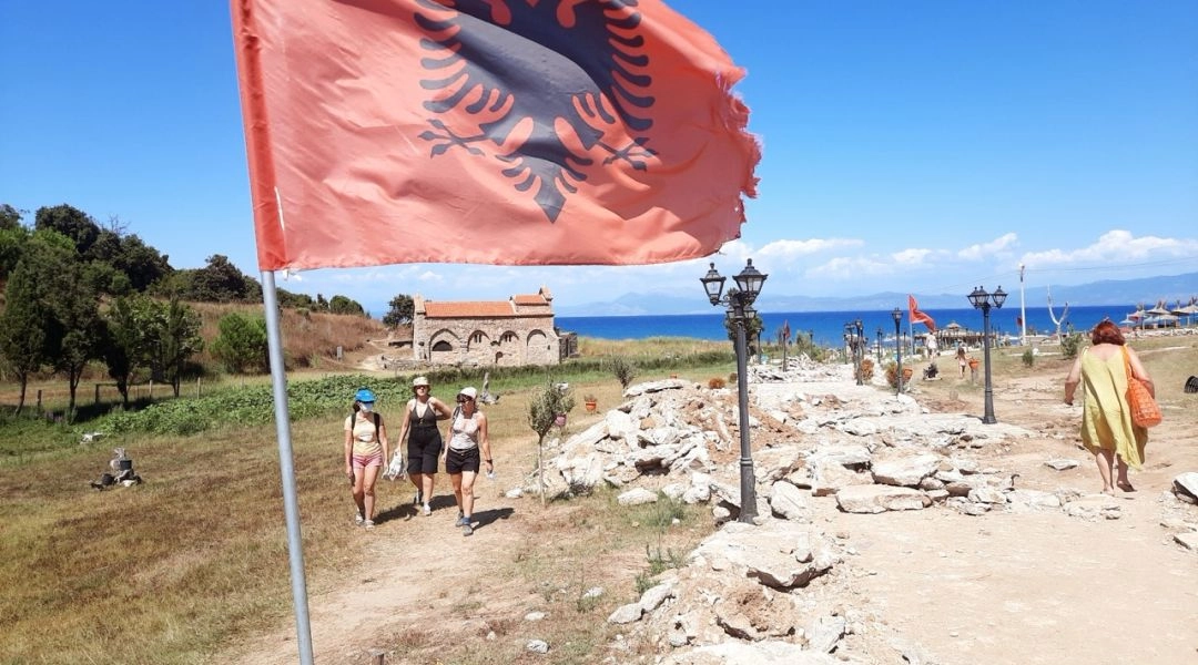 Flaga Albanii z czarnym dwugłowym orłem na tle plaży, morza i kamienistego wybrzeża.