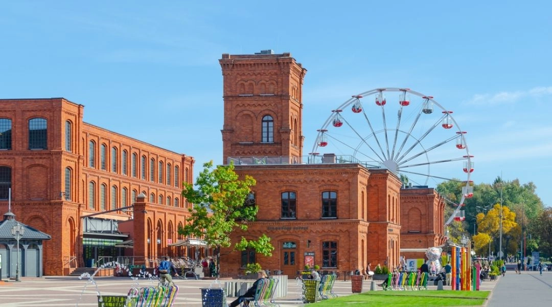 Dziedziniec kompleksu Manufaktura w Łodzi w słoneczny dzień. Na pierwszym planie widać nowoczesną fontannę oraz kolorowe, ażurowe ławki, na których siedzi kilka osób. W głębi wznoszą się majestatyczne, czerwone, ceglane budynki dawnej fabryki, za którymi