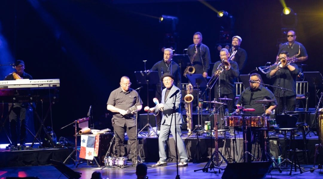 Zespół jazzowy gra koncert na dużej scenie oświetlonej kolorowymi reflektorami.