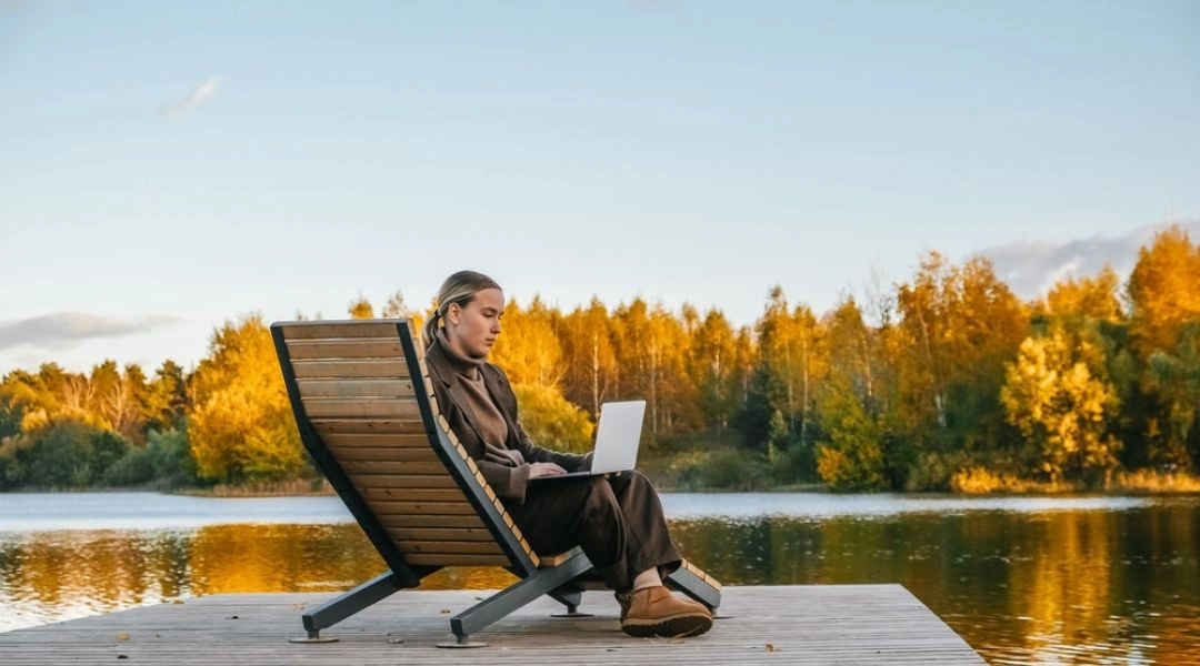 Kobieta pracująca na laptopie na drewnianym pomoście nad jeziorem w otoczeniu jesiennych drzew.