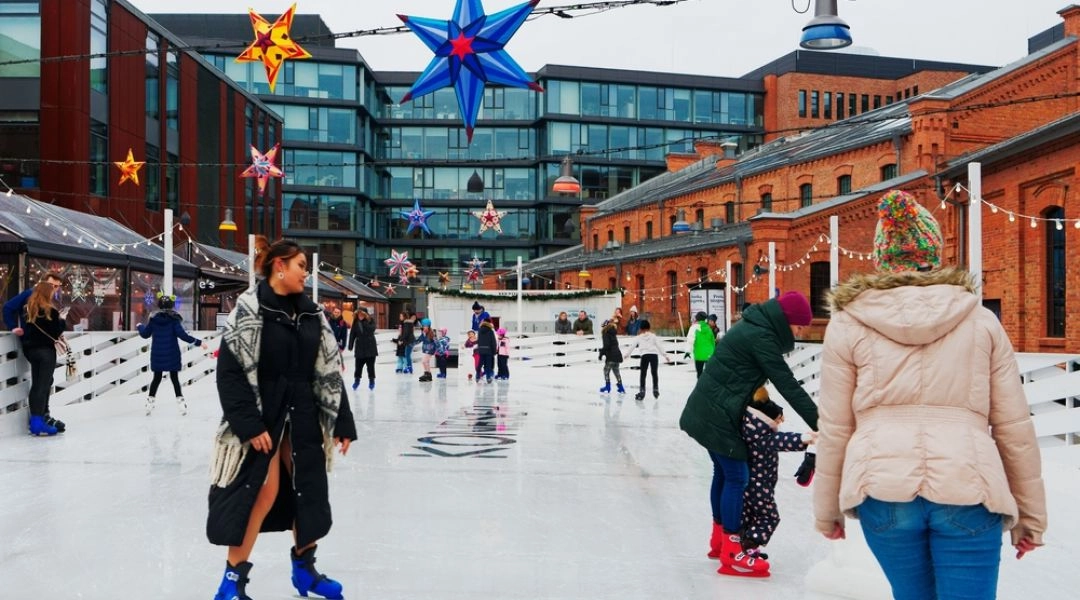 Osoby jeżdżące na łyżwach na lodowisku w Koneserze, otoczone ceglaną zabudową i świątecznymi dekoracjami.