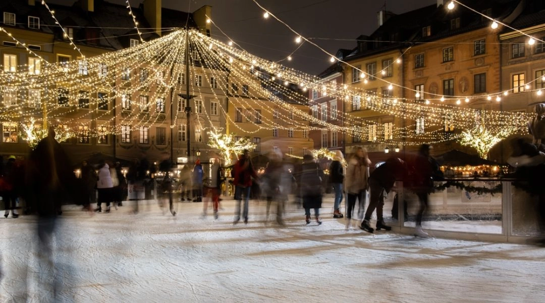 Lodowisko na warszawskiej Starówce oświetlone świątecznymi iluminacjami, z ludźmi jeżdżącymi na łyżwach po zmroku.