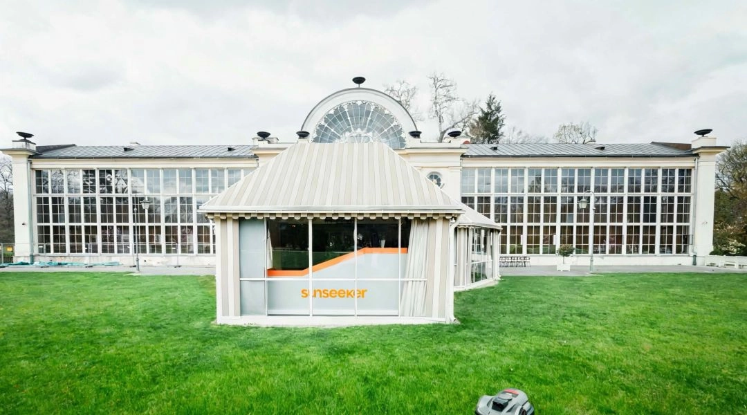 Robot koszący na trawniku przed jasnym pawilonem i dużą przeszkloną oranżerią w ogrodzie