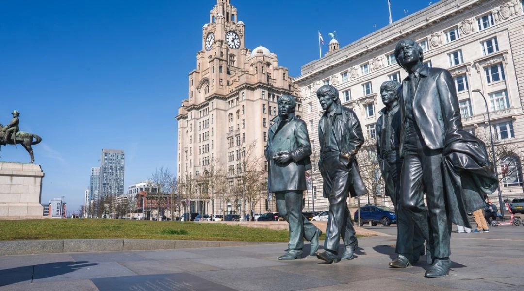 Pomnik członków zespołu The Beatles na nabrzeżu w Liverpoolu, z Royal Liver Building i zabytkowymi budynkami w tle.