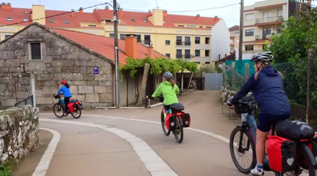 Trzech rowerzystów jadących wzdłuż zabudowań na Camino Portugalskim, z sakwami i kaskami, na wąskiej asfaltowej ścieżce.