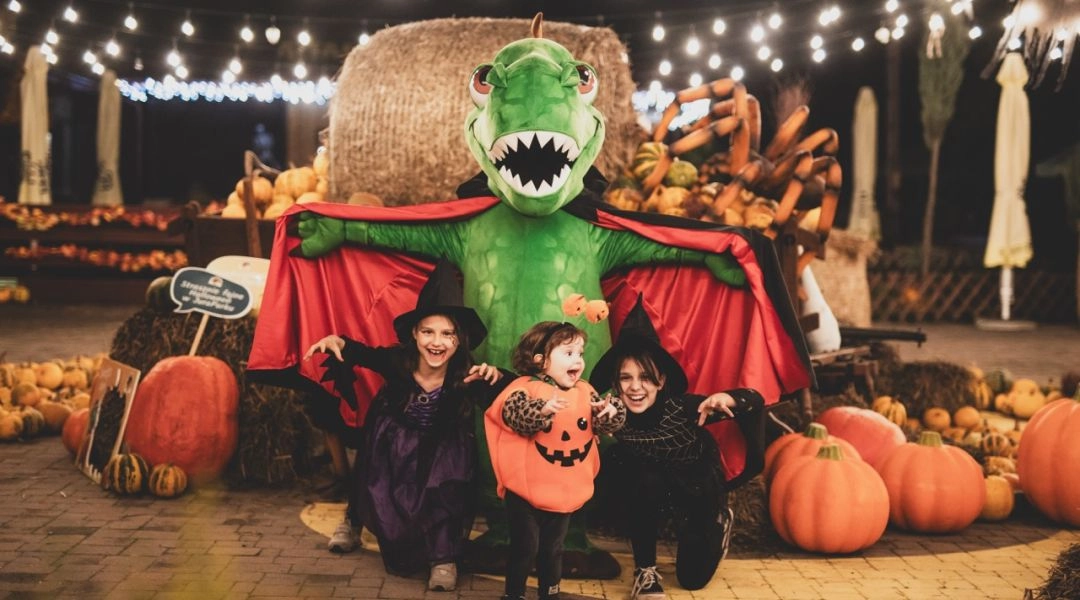 Troje dzieci w halloweenowych przebraniach pozuje z zielonym dinozaurem w pelerynie wśród dyń i jesiennych dekoracji.