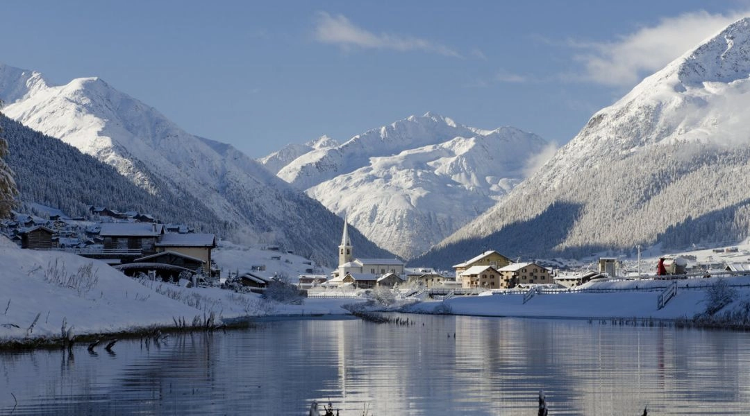 Zimowy widok na miasteczko Livigno z ośnieżonymi górami i kościołem w dolinie, odbijającymi się w spokojnej tafli jeziora.