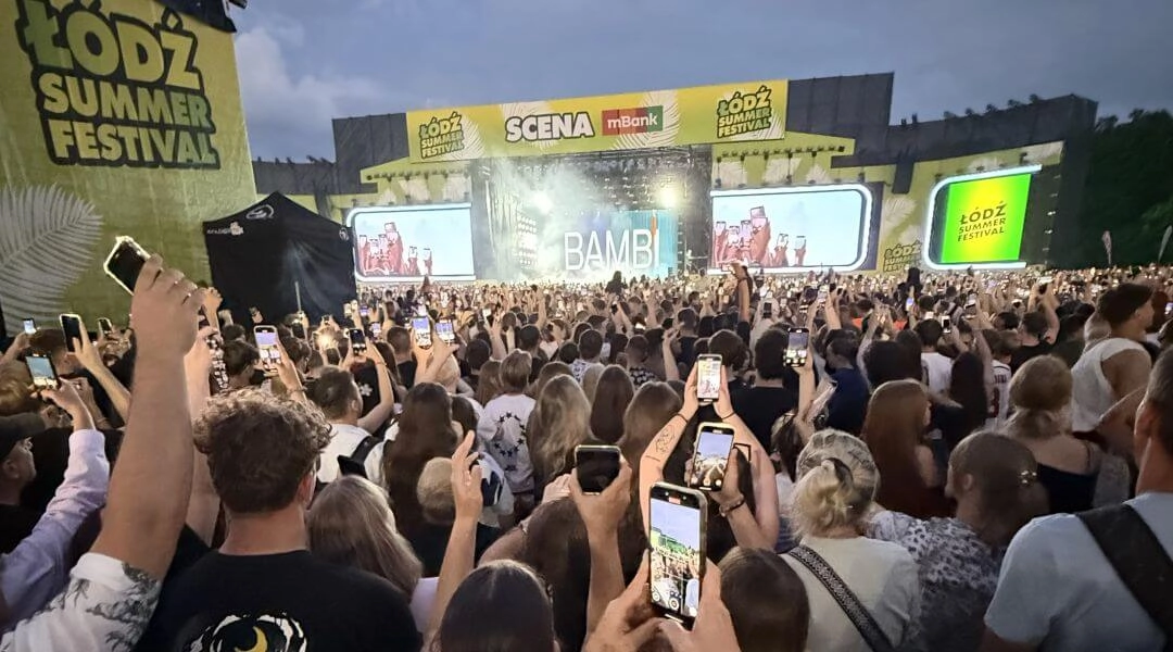 Publiczność z uniesionymi rękami przed sceną Łódź Summer Festival oświetloną czerwonym światłem.