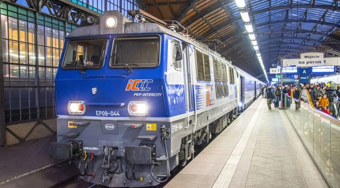 Niebiesko-szara lokomotywa PKP Intercity z wagonami przy peronie pod przeszklonym dachem hali dworcowej.