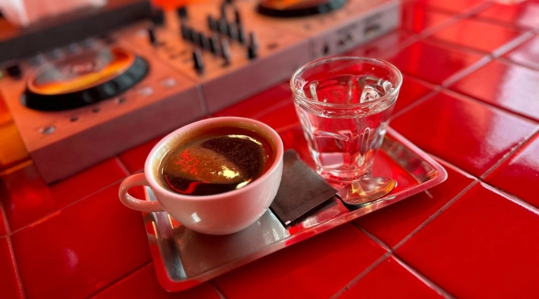Filiżanka espresso i szklanka wody na czerwonym barze, w tle profesjonalny sprzęt DJ-ski.