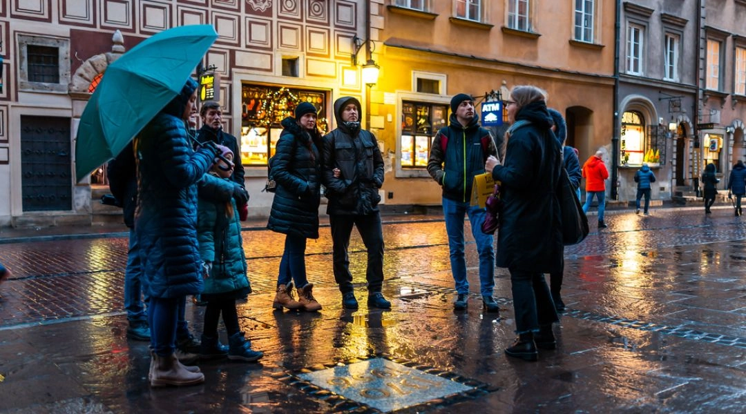Grupa osób słucha przewodniczki podczas spaceru po Starym Mieście w Warszawie, wieczór, mokry bruk, kamienice w tle.