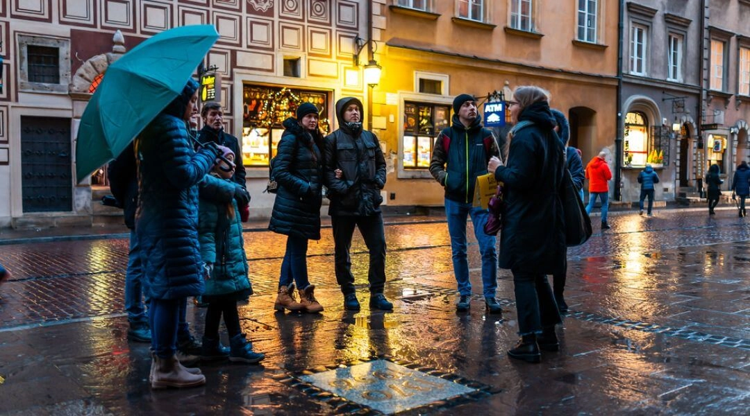 Grupa osób słucha przewodniczki podczas spaceru po Starym Mieście w Warszawie, wieczór, mokry bruk, kamienice w tle.