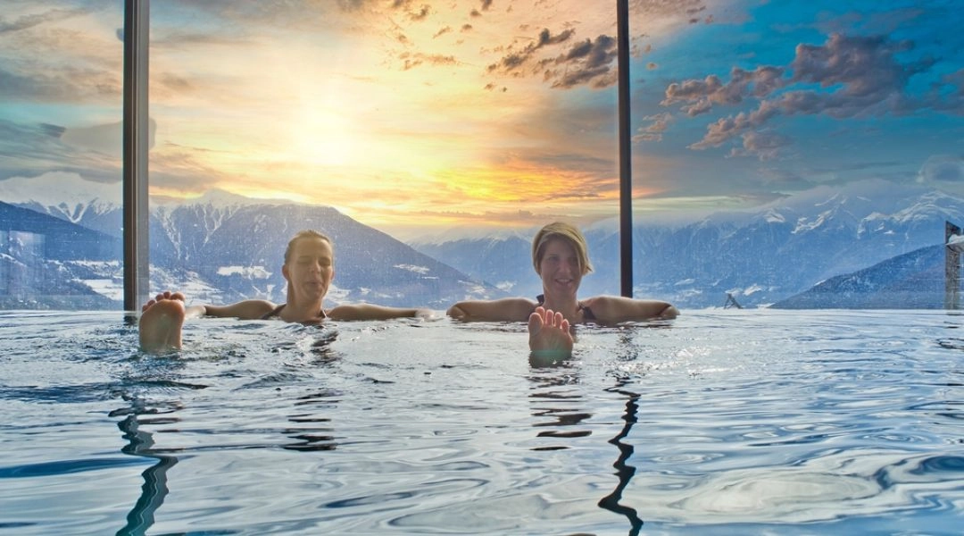 Dwie osoby w podgrzewanym basenie infinity z widokiem na ośnieżone góry o zachodzie słońca.