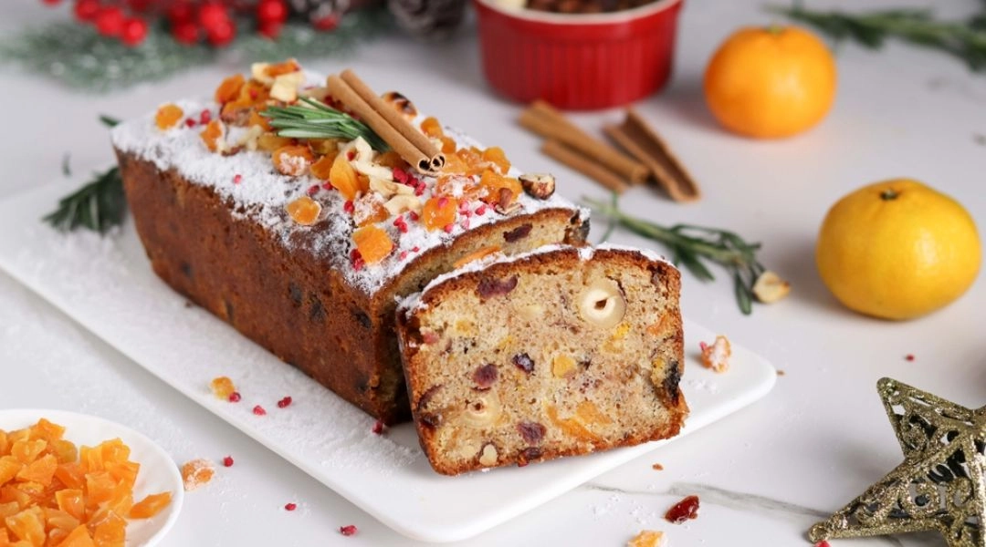 Świąteczny keks z bakaliami na białej paterze, z widocznymi kawałkami orzechów, suszonych owoców i skórki pomarańczowej, posypany cukrem pudrem, obok przypraw korzennych i cytrusów.