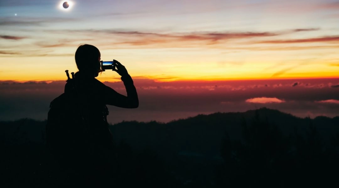 Osoba fotografuje zaćmienie Słońca telefonem na tle gór i zachodzącego nieba.