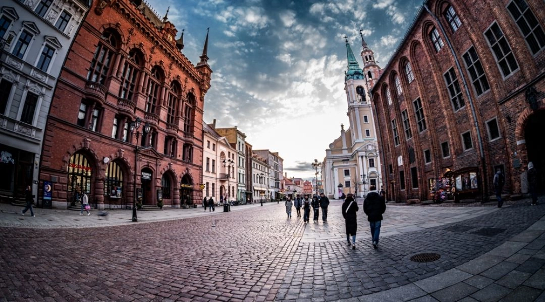 Brukowany Rynek Staromiejski w Toruniu z ceglanymi zabytkowymi budynkami, kościołem i spacerującymi ludźmi.