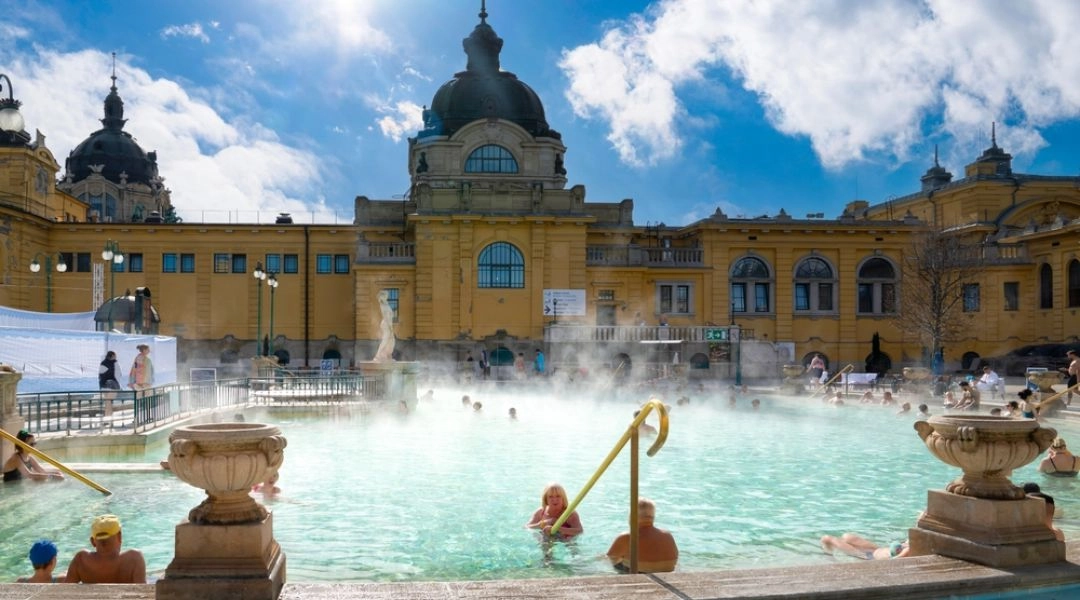 Odkryty basen termalny kąpieliska Széchenyi w Budapeszcie. Nad wodą unosi się para, a w tle widać charakterystyczny żółty budynek z kopułą i osoby korzystające z kąpieli.