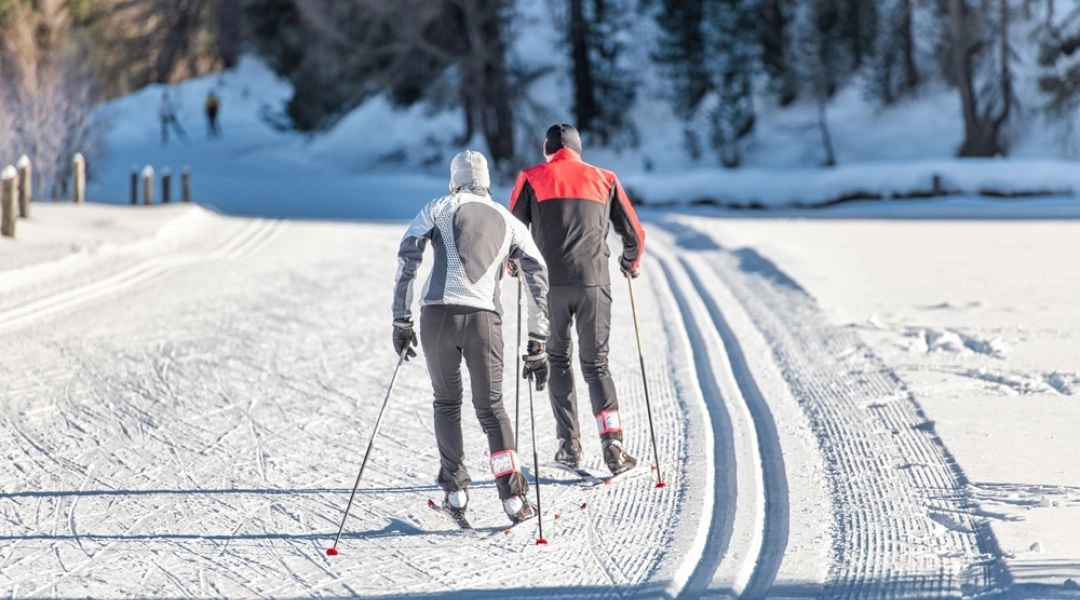 Dwie osoby biegnące na nartach biegowych po przygotowanym śladzie w zimowym lesie.