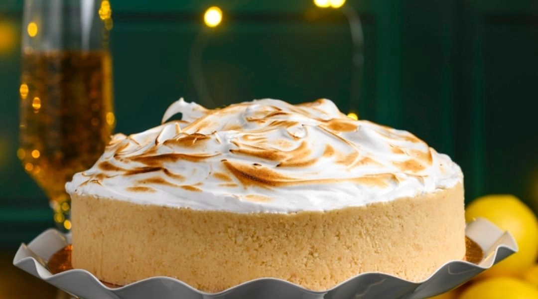 Świąteczny sernik z rumianą bezą na białej paterze, ustawiony na stole z cytrynami, kieliszkiem prosecco i złotymi światełkami w tle, w eleganckiej bożonarodzeniowej aranżacji.