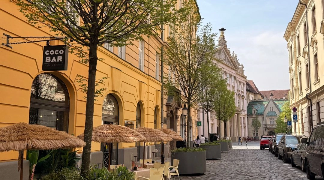 Brukowana ulica w centrum Bratysławy z ogródkiem kawiarni, jasnymi kamienicami i drzewami przy fasadach.