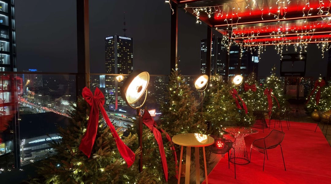 Panorama nocnej Warszawy widziana z tarasu Winter WOWderland, z oświetlonymi choinkami i dekoracjami w pierwszym planie.