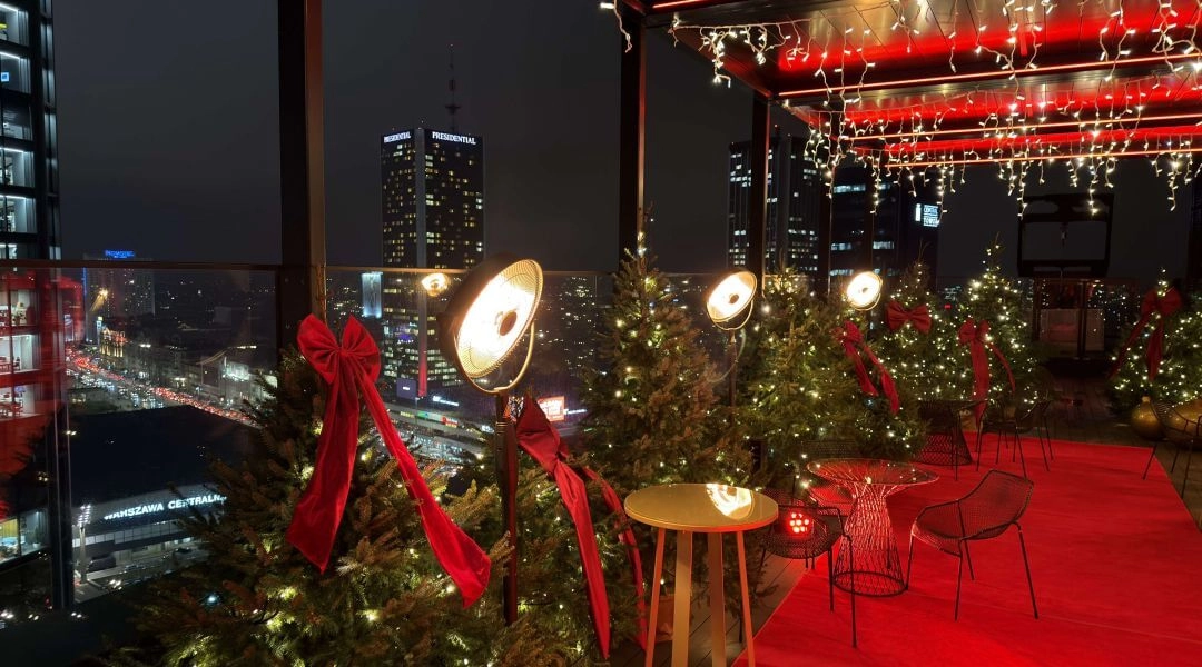 Panorama nocnej Warszawy widziana z tarasu Winter WOWderland, z oświetlonymi choinkami i dekoracjami w pierwszym planie.