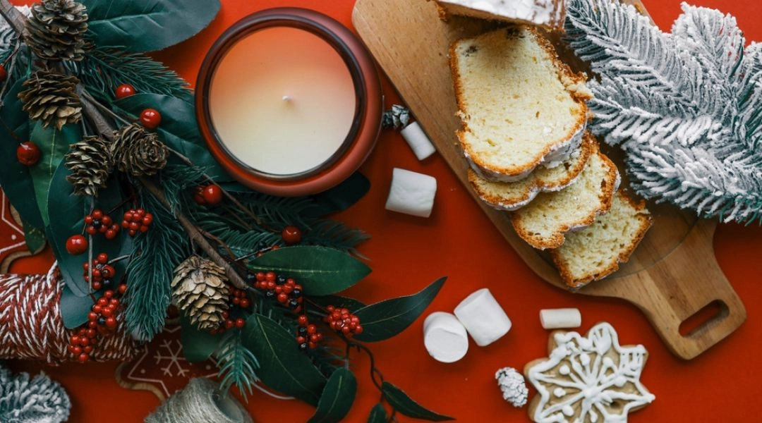 Świąteczna drożdżówka posypana cukrem pudrem, pokrojona na plastry na drewnianej desce, obok świeca, gałązki świerku, pierniczki i zimowe dekoracje na czerwonym tle.