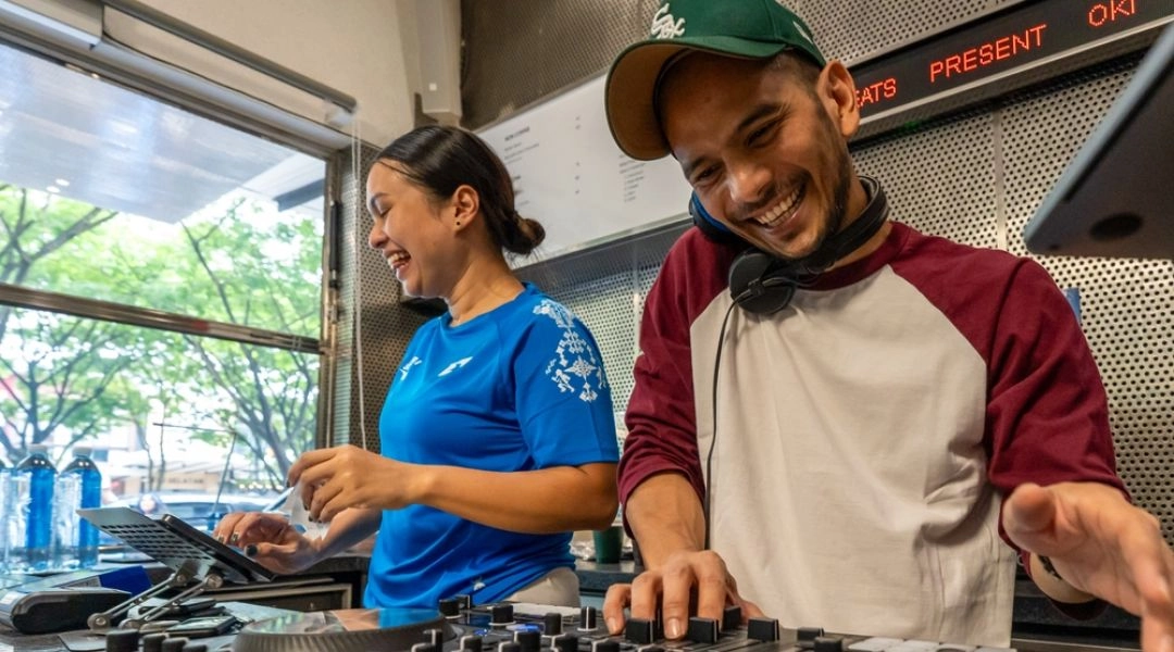 Uśmiechnięty DJ miksujący muzykę przy konsoli w kawiarni, obok kobieta przygotowująca zamówienia.