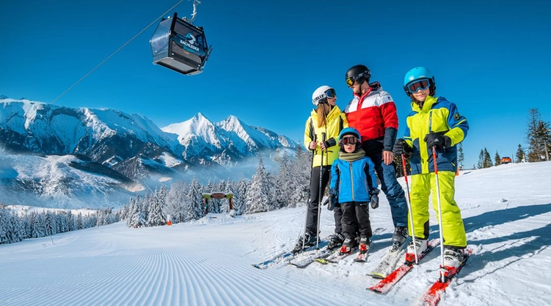 Rodzina z dziećmi na nartach w ośrodku Bachledka Ski & Sun, stoją na przygotowanym stoku, nad nimi gondola i widok na Tatry.