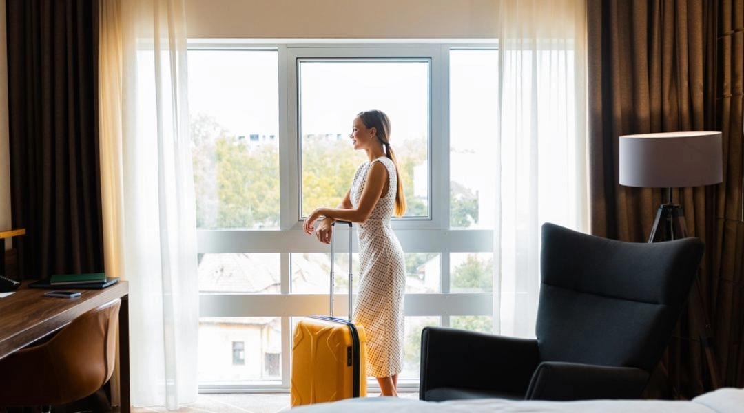 Kobieta z walizką stojąca przy oknie w pokoju hotelowym