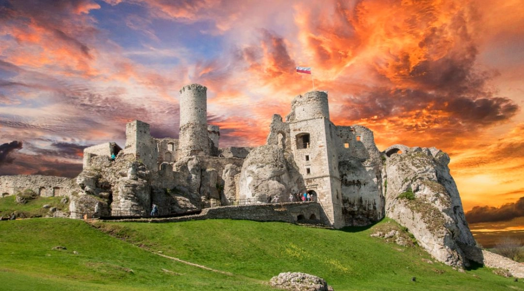 Zamek Ogrodzieniec na wzgórzu o zachodzie słońca, kamienne ruiny i zielone wzgórza wokół.