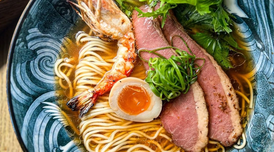 Ramen w ozdobnej niebieskiej misce z krewetką, plastrami mięsa, jajkiem, makaronem, szczypiorkiem i zielonymi liśćmi.