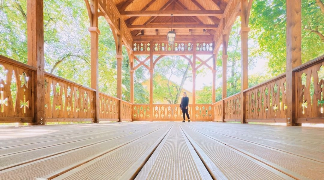 Długa drewniana weranda z ozdobnymi balustradami, wysokim dachem i osobą stojącą na jej końcu.