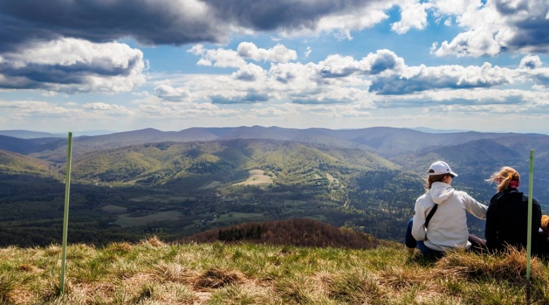 Panorama Bieszczad z punktu widokowego, para siedząca na trawie i rozległe zielone wzgórza pod chmurami.