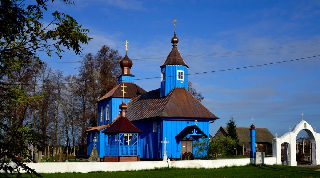 Niebieska drewniana cerkiew z kopułami na Podlasiu, otoczona zielenią i cmentarzem przykościelnym.