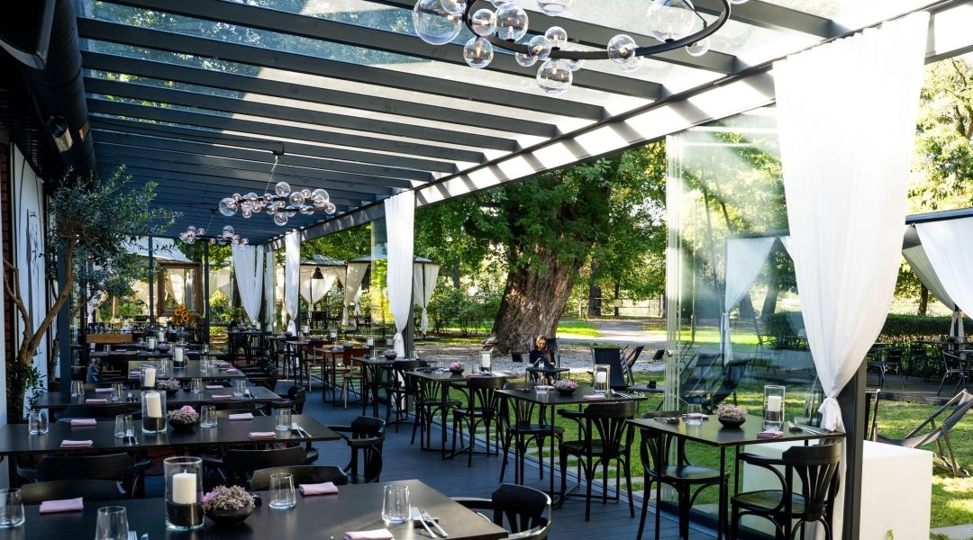 Oranżeria restauracji KLONN w Warszawie, czarne stoliki pod szklanym dachem i widok na ogród ze starym drzewem.