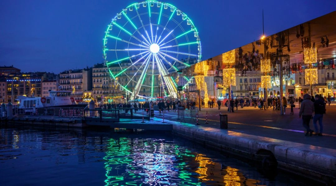 Vieux-Port w Marsylii po zmroku, z dużym diabelskim młynem i świątecznymi iluminacjami odbijającymi się w wodzie.