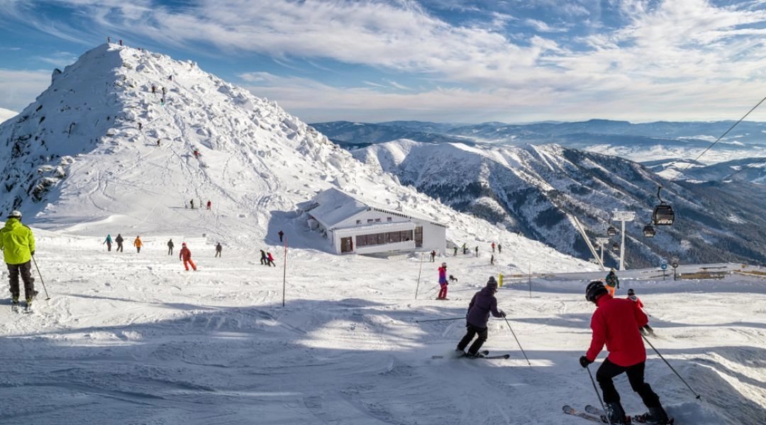 Narciarze jadący szeroką trasą w ośrodku Jasná w Niskich Tatrach, w tle zaśnieżone szczyty i górna stacja kolei linowej.