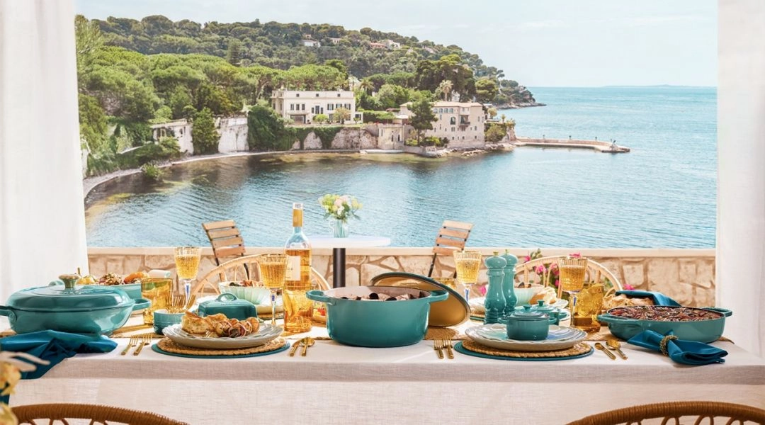 Stół z naczyniami Le Creuset w kolorze Riviera Bleu ustawiony przy widoku na morze i śródziemnomorskie wybrzeże.