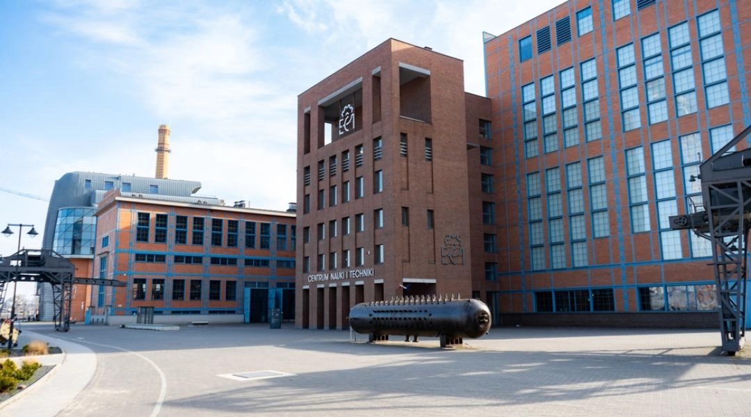 Widok z zewnątrz na nowoczesny, ceglany budynek Centrum Nauki i Techniki EC1 w Łodzi z napisem na fasadzie. Przed wejściem stoi duża, ciemna instalacja techniczna w kształcie cylindra, a obok widać fragment dawnej infrastruktury przemysłowej. Nad kompleks
