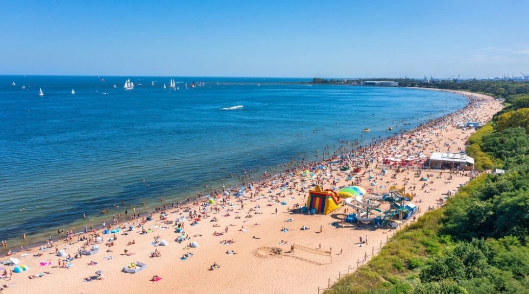 Plaża w gdańskim Brzeźnie widziana z góry, z szerokim pasem piasku, turystami odpoczywającymi nad wodą i zielenią przy brzegu.
