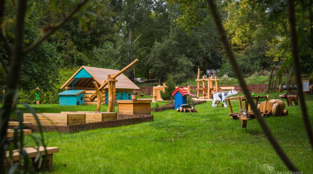 Naturalny plac zabaw w Dolince Służewskiej w Warszawie z drewnianymi domkami, figurami zwierząt i zielonym otoczeniem.