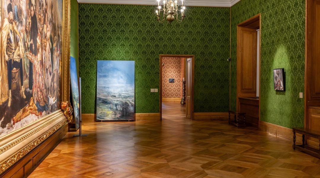 Zielona sala Zamku Królewskiego w Warszawie z obrazami Łukasza Stokłosy ustawionymi obok monumentalnego obrazu historycznego, parkiet z jasnego drewna i klasyczne oświetlenie.